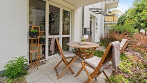 Un espace extérieur aménagé avec une table ronde et deux chaises en bois, un présentoir à plantes en bois et une porte vitrée donnant sur un balcon.