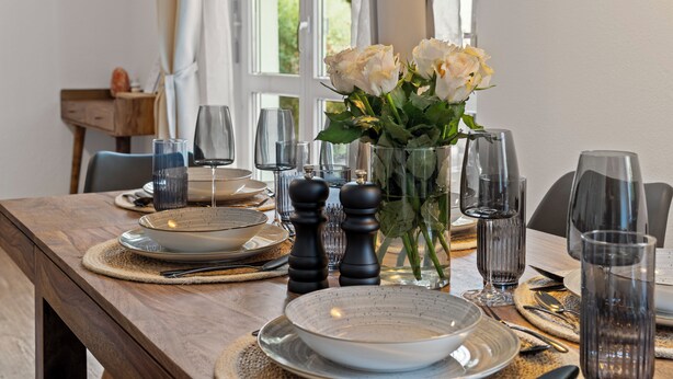 Une table à manger dressée avec des assiettes, des bols, des couverts, des verres et un vase rempli de fleurs.