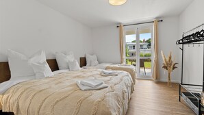 Une chambre d’hôtel avec deux lits, un plancher en bois, une grande fenêtre donnant sur une maison, et un porte-manteau noir sur lequel sont suspendus des vêtements.