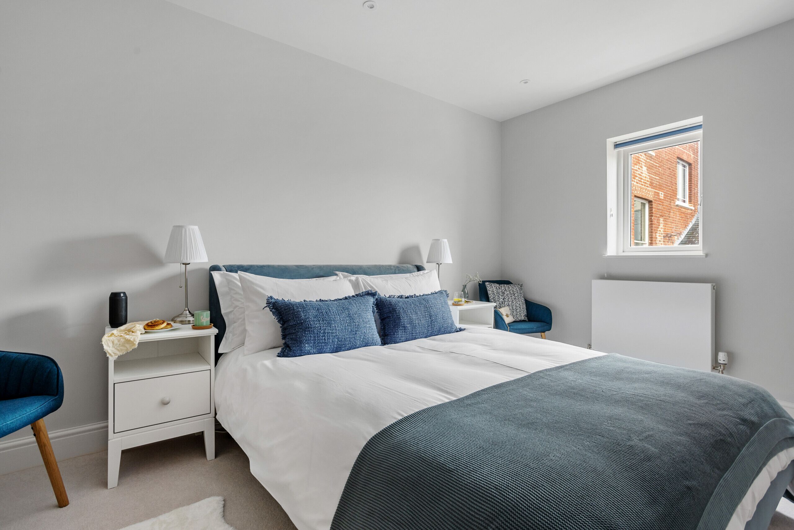 A bedroom with a bed, nightstands, a lamp, a chair, and a window with a view of a brick building.