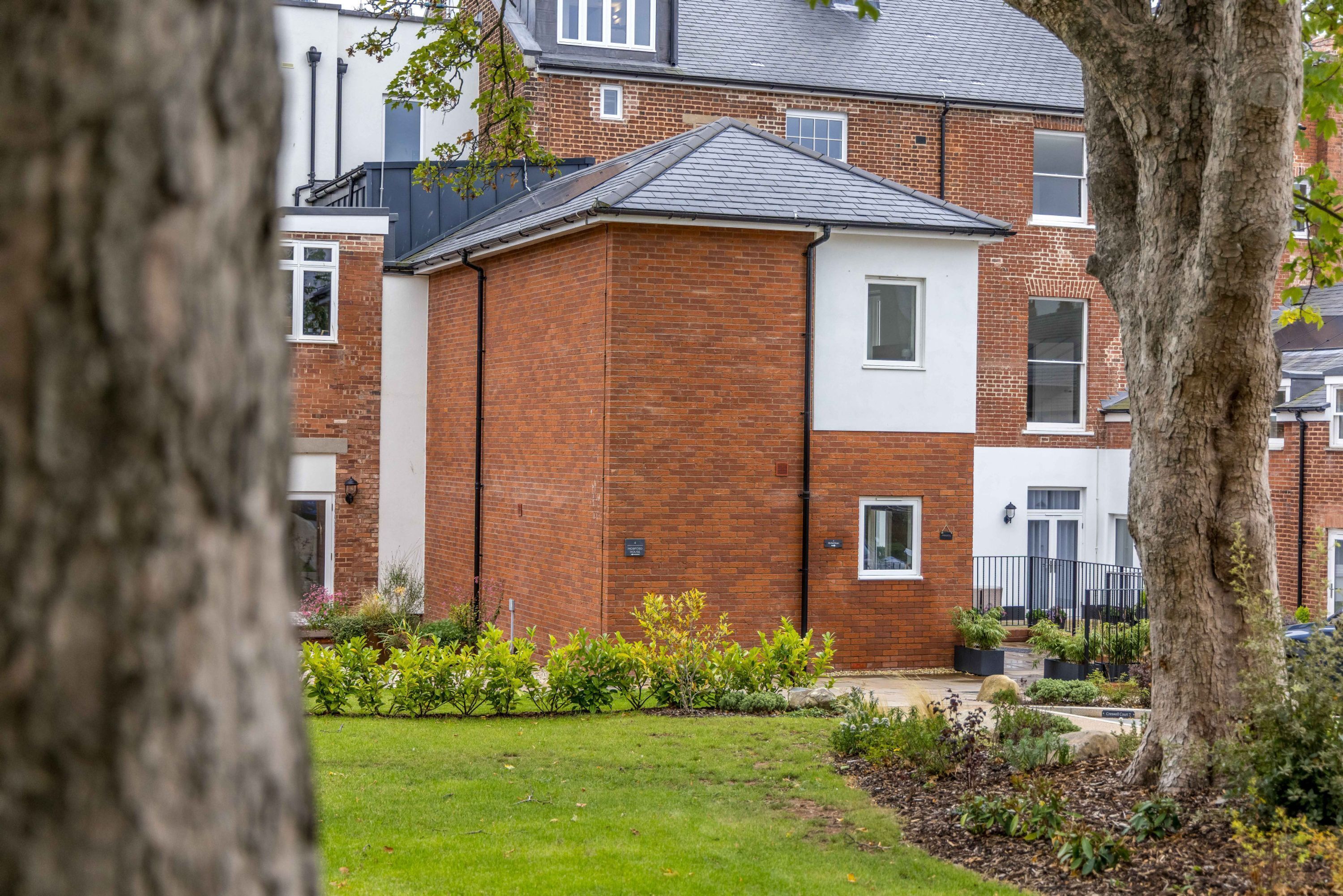 Un immeuble résidentiel à la façade en briques, doté d’un toit plat et d’un petit jardin devant, agrémenté d’arbustes et d’une pelouse.