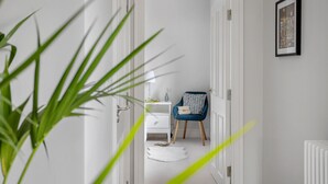 A hallway with a white door, a blue chair with a patterned cushion, a white nightstand with a vase, and a framed picture on the wall. - Morford House in Exeter (Exeter)