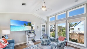 Een ruime woonkamer met een grote televisie waarop een strandscène wordt getoond, een plafondventilator en uitzicht op de achtertuin.