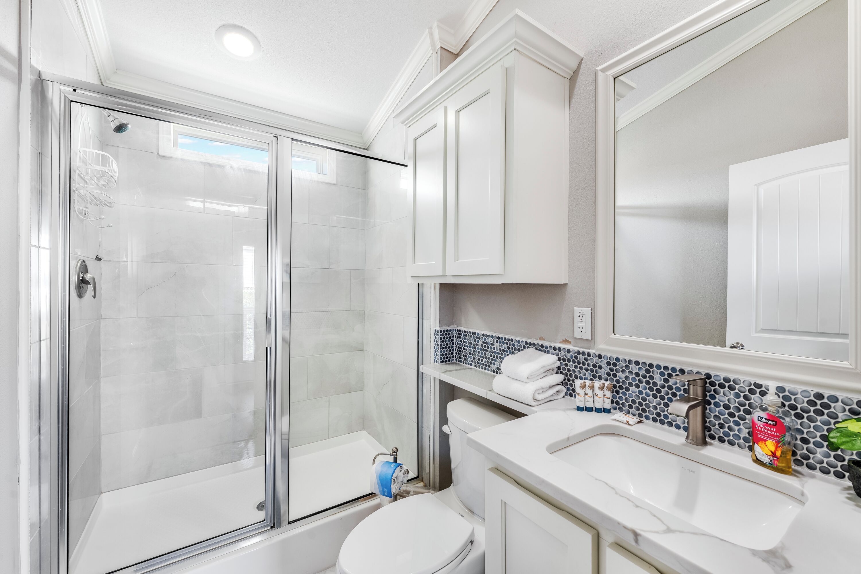 Un baño moderno con ducha envuelta en vidrio, inodoro blanco, lavamanos con espejo y salpicadero de azulejos.
