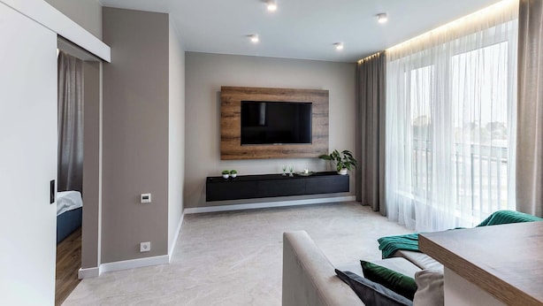 Una sala moderna con un televisor de pantalla plana montado en un panel de madera, un mueble de entretenimiento negro debajo y un gran ventanal con cortinas translúcidas.