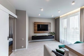 Uma sala de estar moderna com uma TV de tela plana presa a um painel de madeira, um móvel de entretenimento preto abaixo e uma janela grande com cortinas finas.