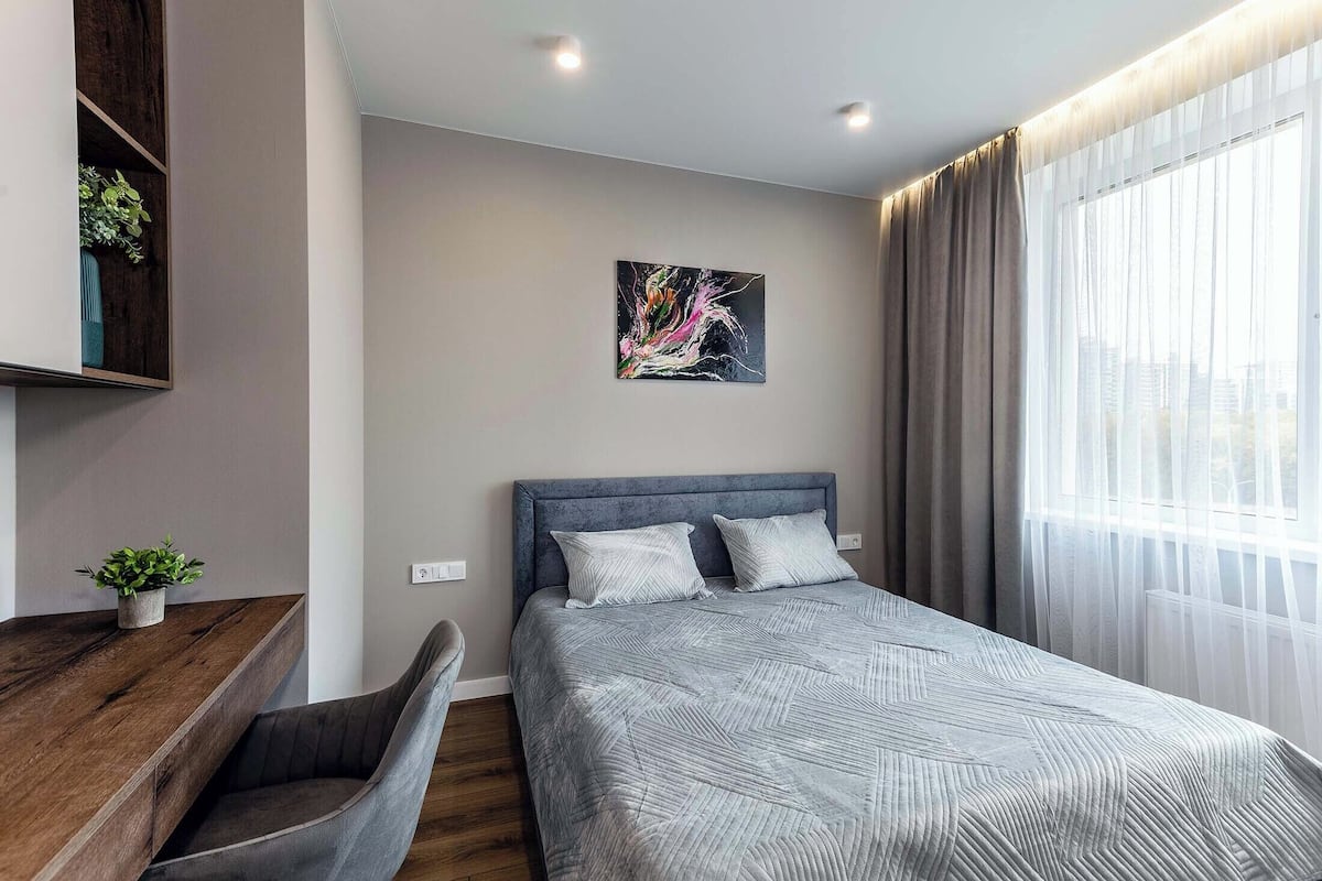Quarto de hotel moderno com uma cama grande, uma mesa com uma planta, um quadro na parede e vista da cidade através de cortinas translúcidas.
