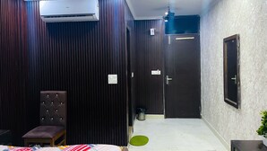A hotel room with a dark wooden panel wall, a single bed with a colorful striped bedspread, a brown upholstered chair, a small green mat, and a ceiling fan. - Roomshala Hotel Tom & Jerry (New Delhi)