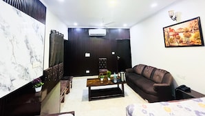 A living room with a dark brown sofa, a wooden coffee table, a marble accent wall, a ceiling fan, and a painting on the wall. - Roomshala Hotel Tom & Jerry (New Delhi)