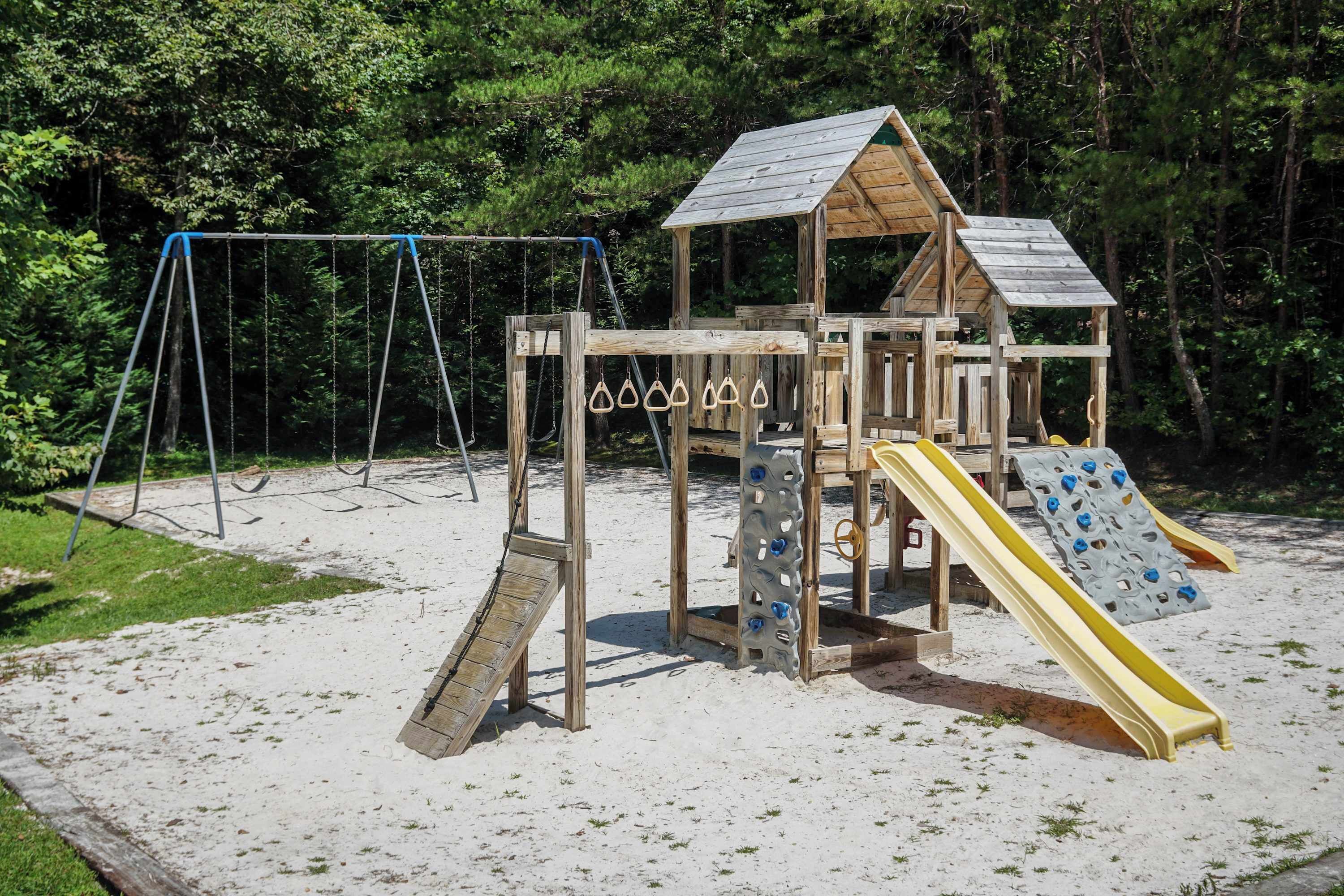 A wooden play structure with a slide and swings, set on sandy ground with trees in the background.