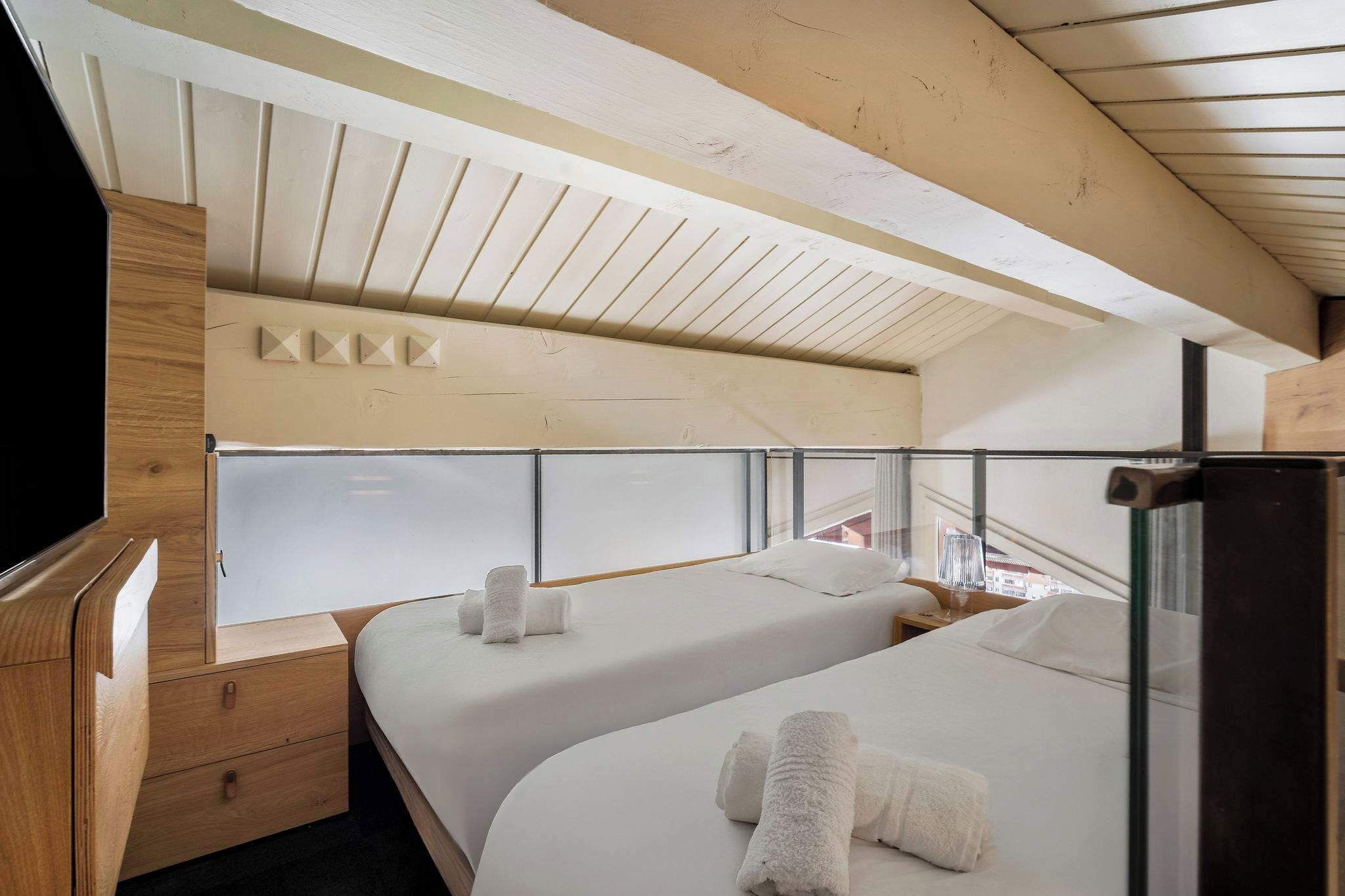 A modern hotel room with two beds, wooden headboard, a nightstand, and a large window offering a city view.