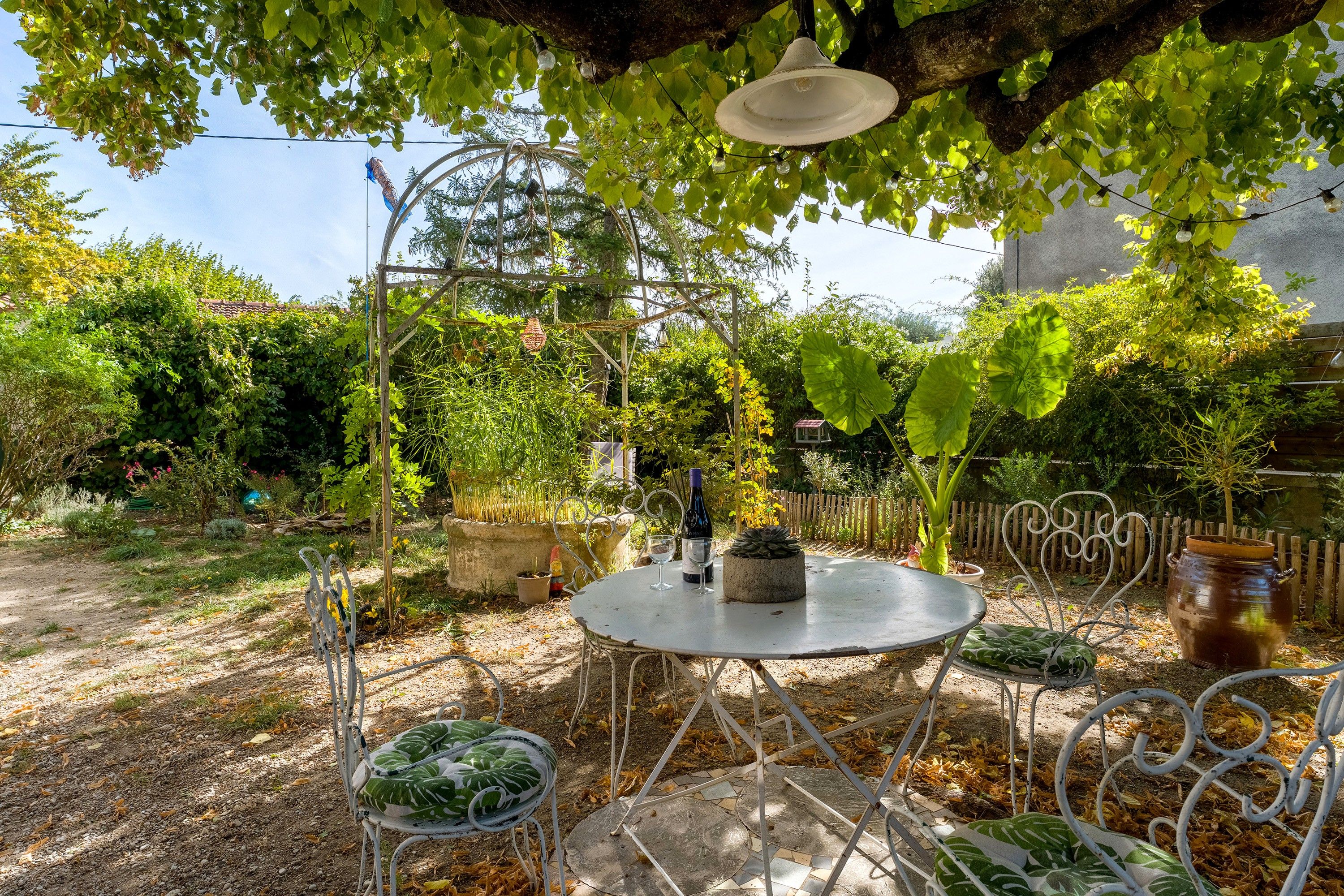Une terrasse de jardin avec une table ronde et des chaises, entourée de verdure et d’une clôture en bois.