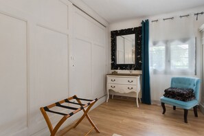 A room with a wooden floor, a white dresser, a mirror with a black frame, a blue chair, and a drying rack. - Charming Apartment - Under the Linden Tree (Valréas)