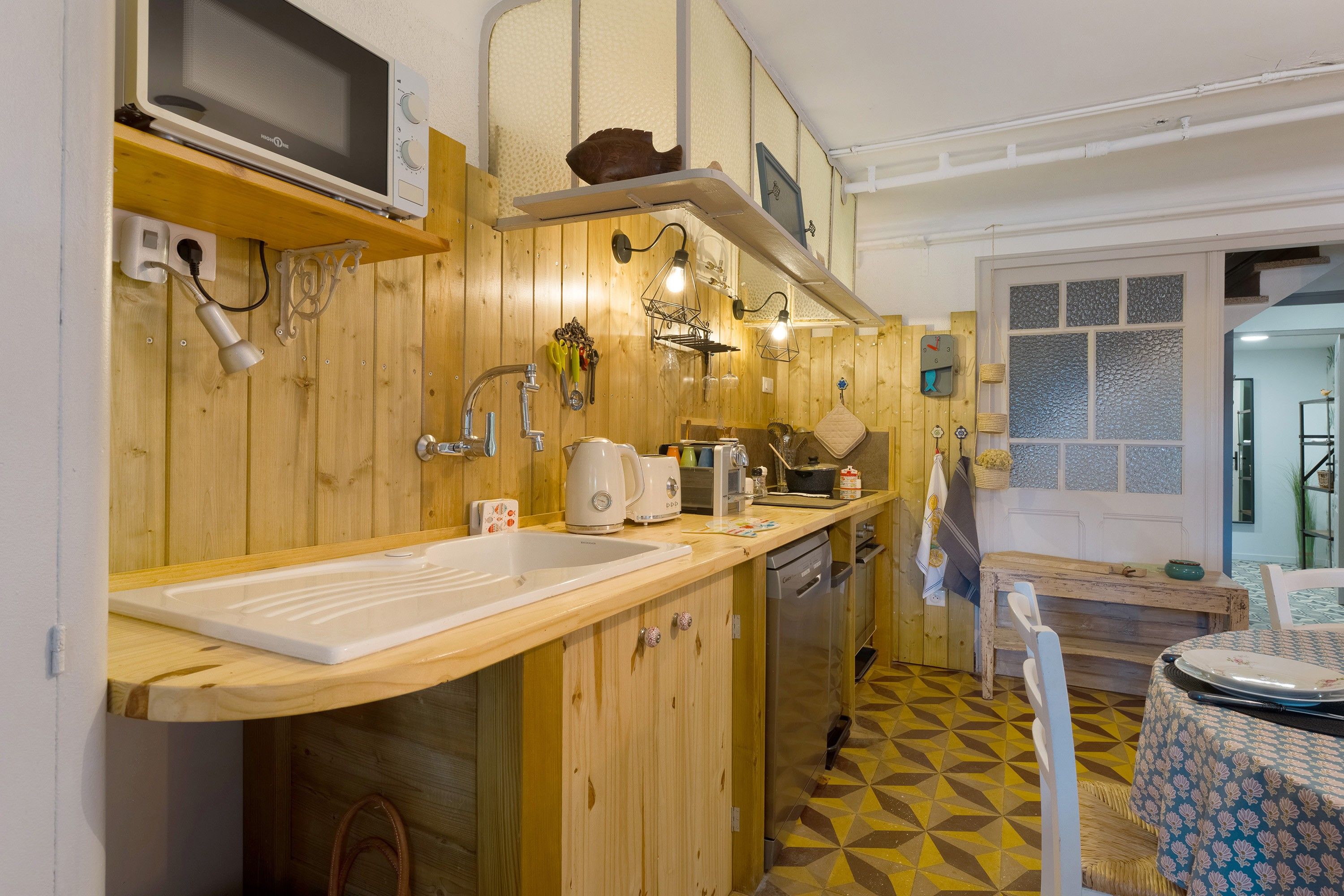 Une cuisine avec des meubles de rangement en bois, un four à micro-ondes, un évier et un coin repas agrémenté d’une nappe à motifs.