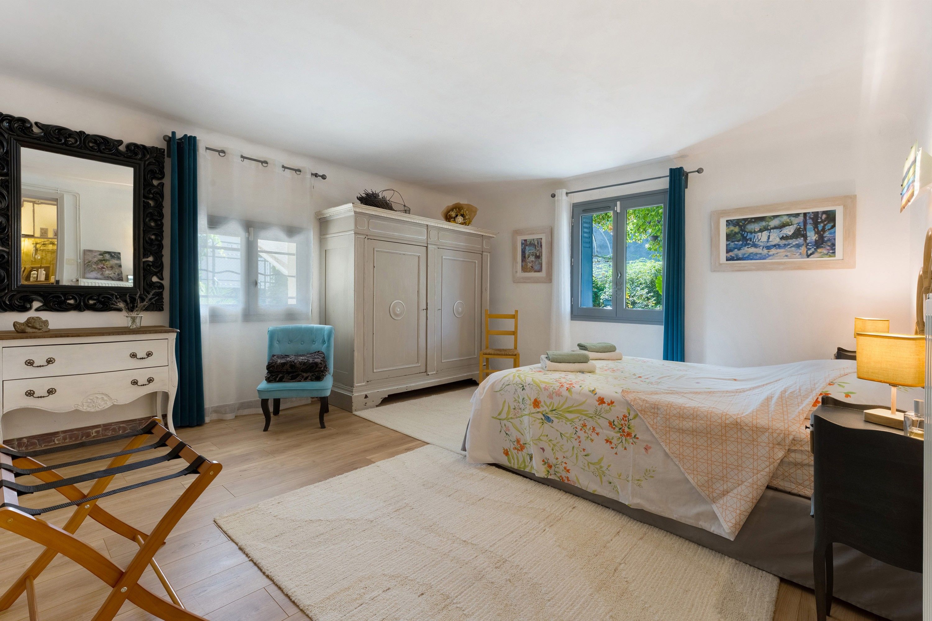 Une chambre à coucher avec un lit, une chaise, une armoire, un miroir et une fenêtre avec des rideaux.