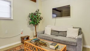 Un salon avec un canapé gris, une table basse en verre, une petite table d’appoint en bois, une grande plante en pot et un téléviseur à écran plat fixé au mur.