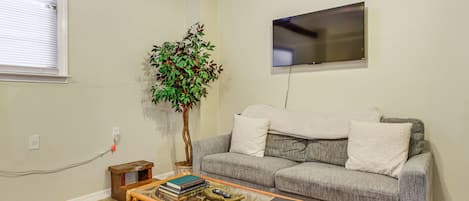 A living room with a grey sofa, a glass coffee table, a small wooden side table, a large potted plant, and a flat-screen TV mounted on the wall.