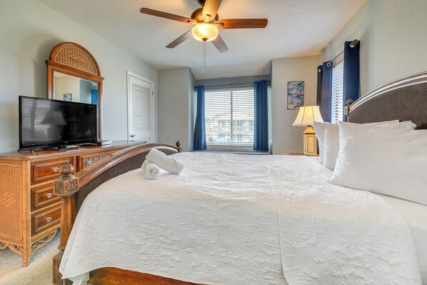A hotel room with a large bed, a television on a wooden dresser, a ceiling fan, and a window with blue curtains.