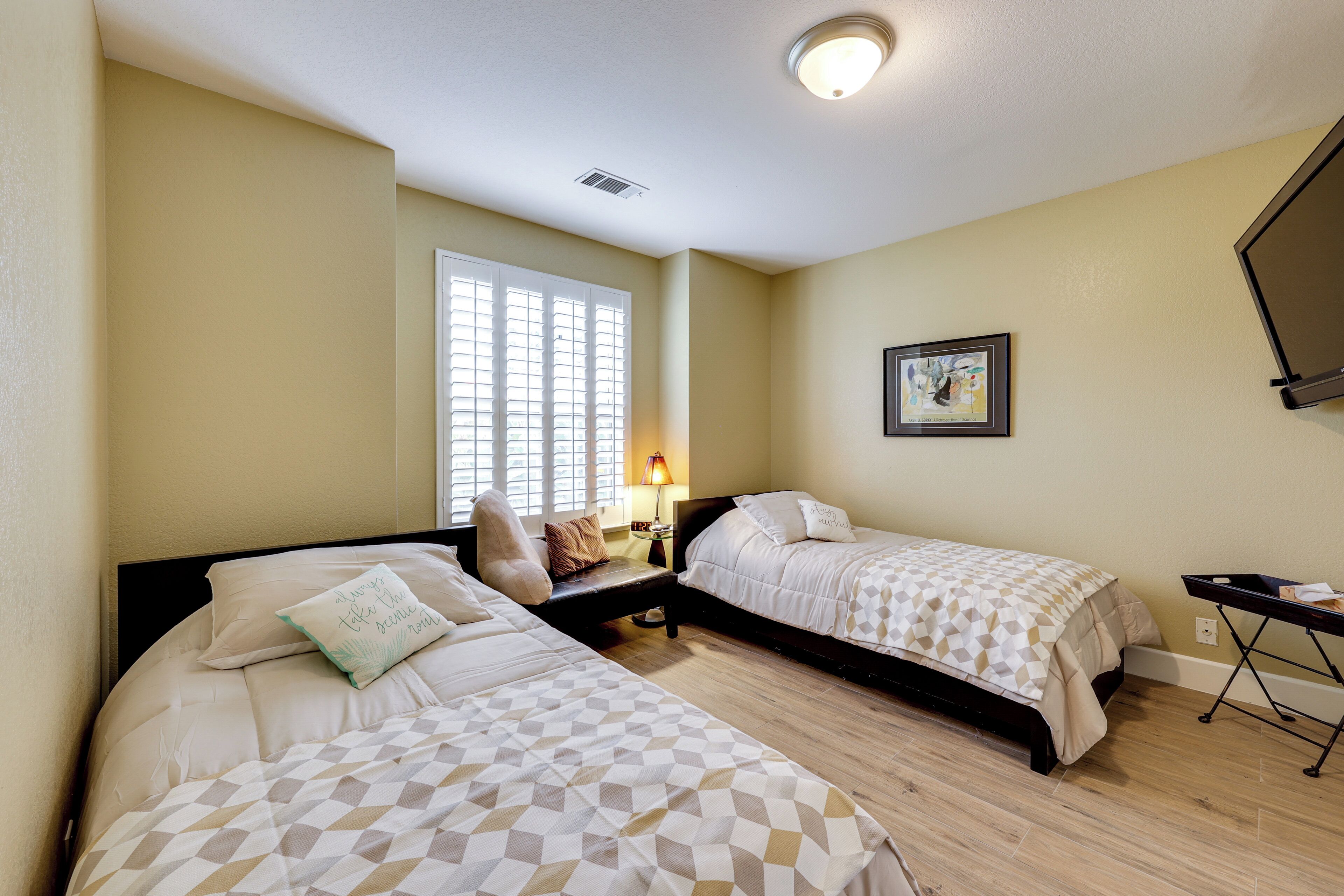 Une chambre d’hôtel avec deux lits, une fenêtre avec des stores, une table de chevet avec une lampe et un téléviseur à écran plat fixé au mur.