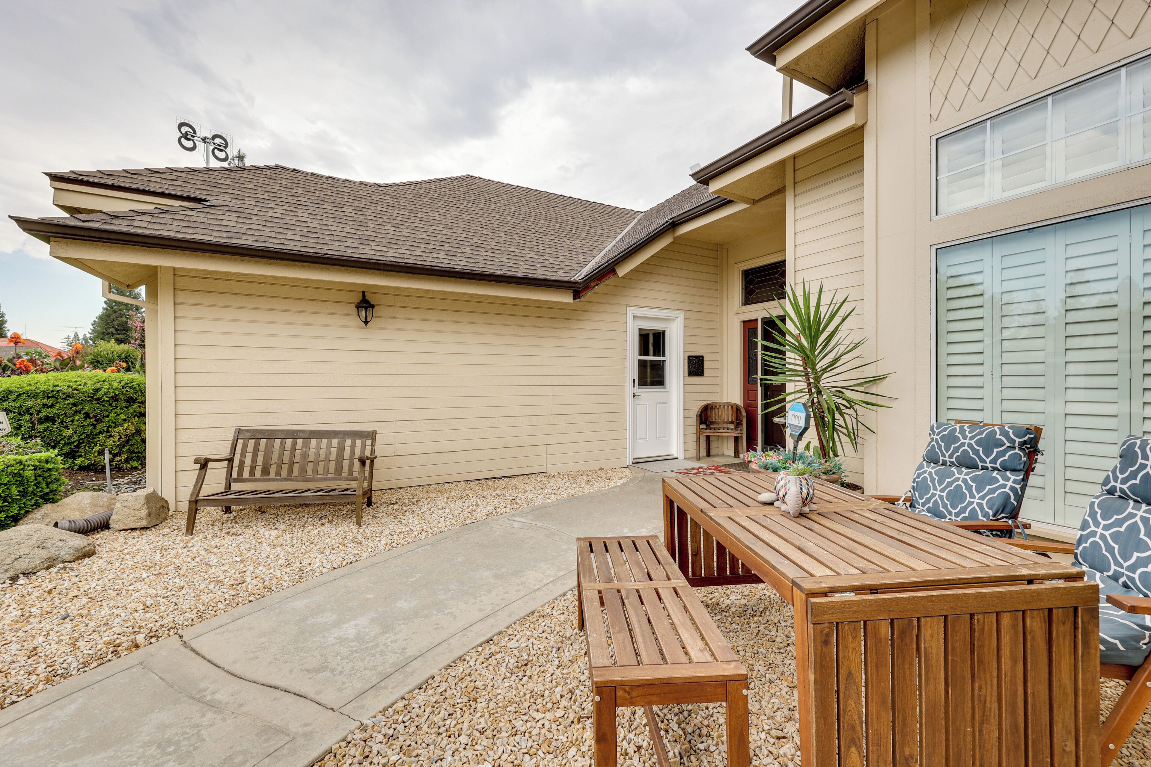 A beige house with a covered patio, a wooden bench, a table, and two patterned chairs.