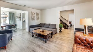 Un vaste salon comprenant un canapé d’angle gris, une table basse en bois et un fauteuil en cuir. On aperçoit à l’arrière-plan un coin repas avec une table et des chaises.