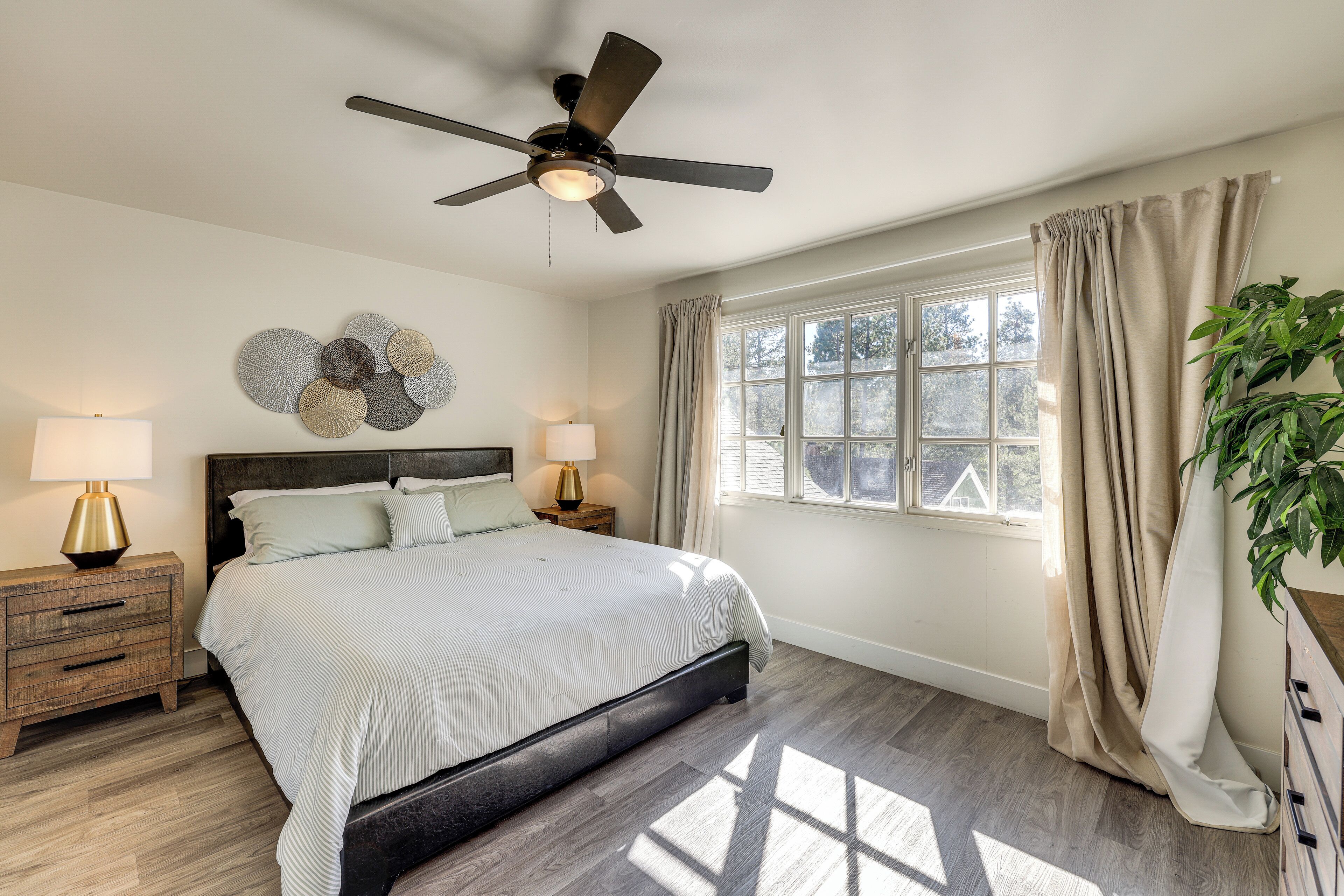 Une chambre à coucher comprenant un lit, des tables de chevet, un ventilateur de plafond, une fenêtre avec des rideaux et une plante.