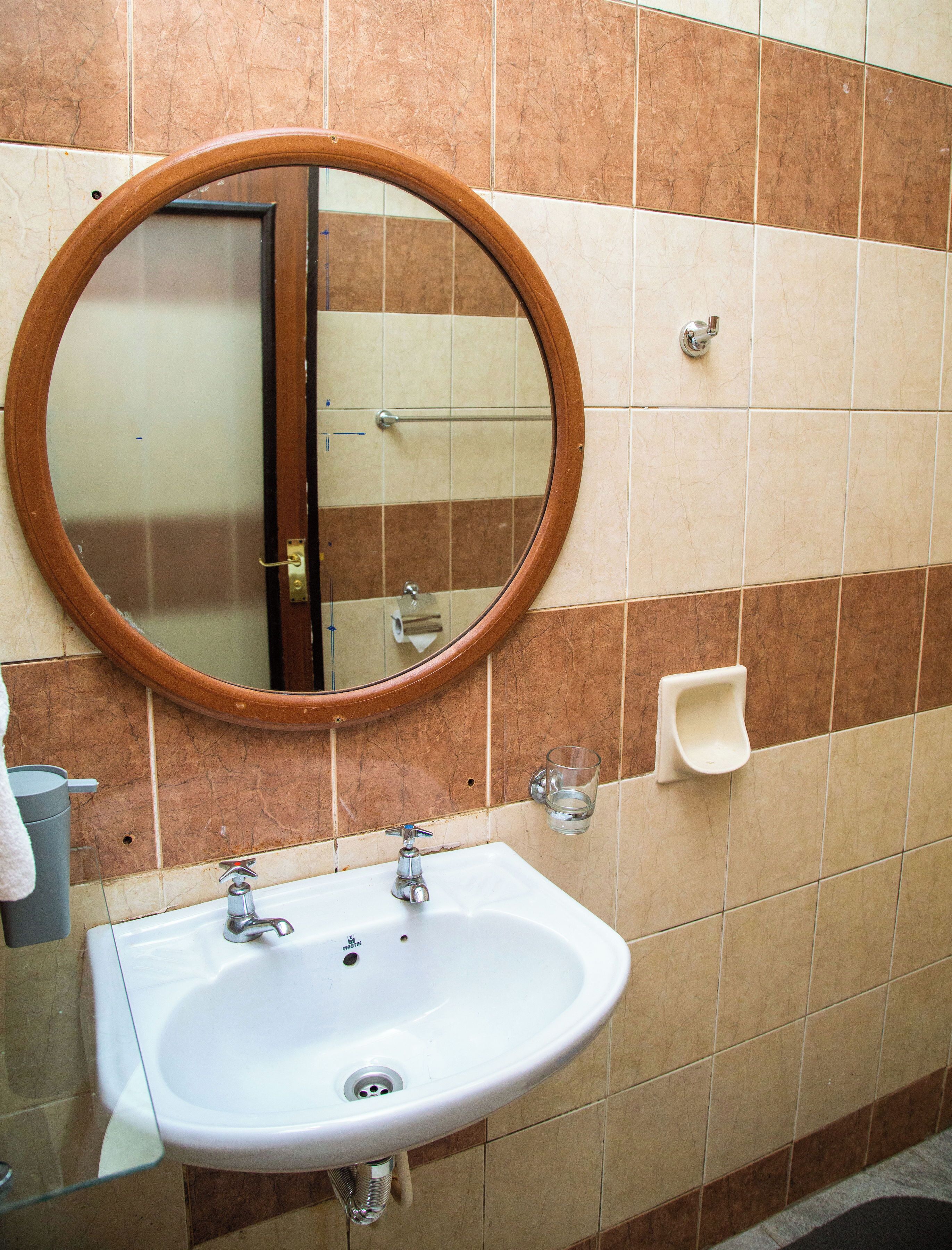 Une salle de bain avec un miroir rond, un lavabo, un distributeur de serviettes et un verre d’eau fixé au mur.