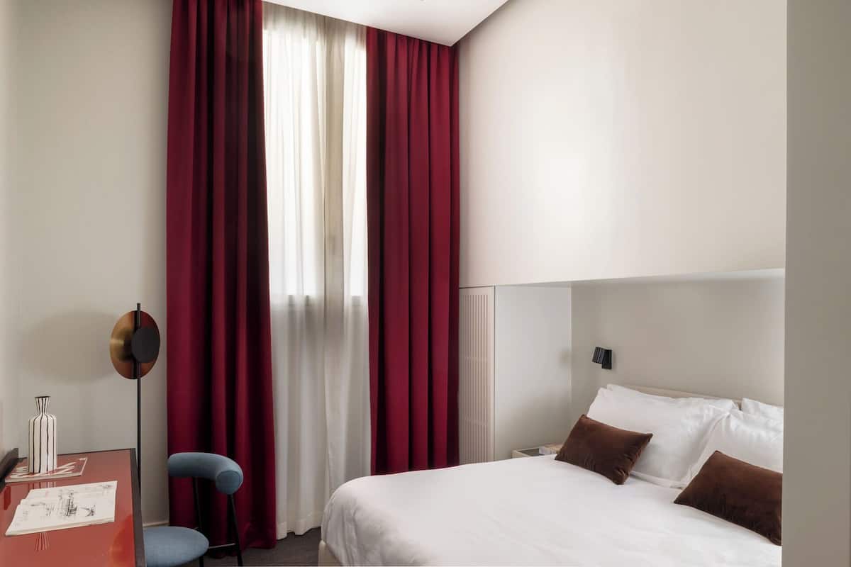 A hotel room with a bed, a desk with a chair, and a window with red curtains.