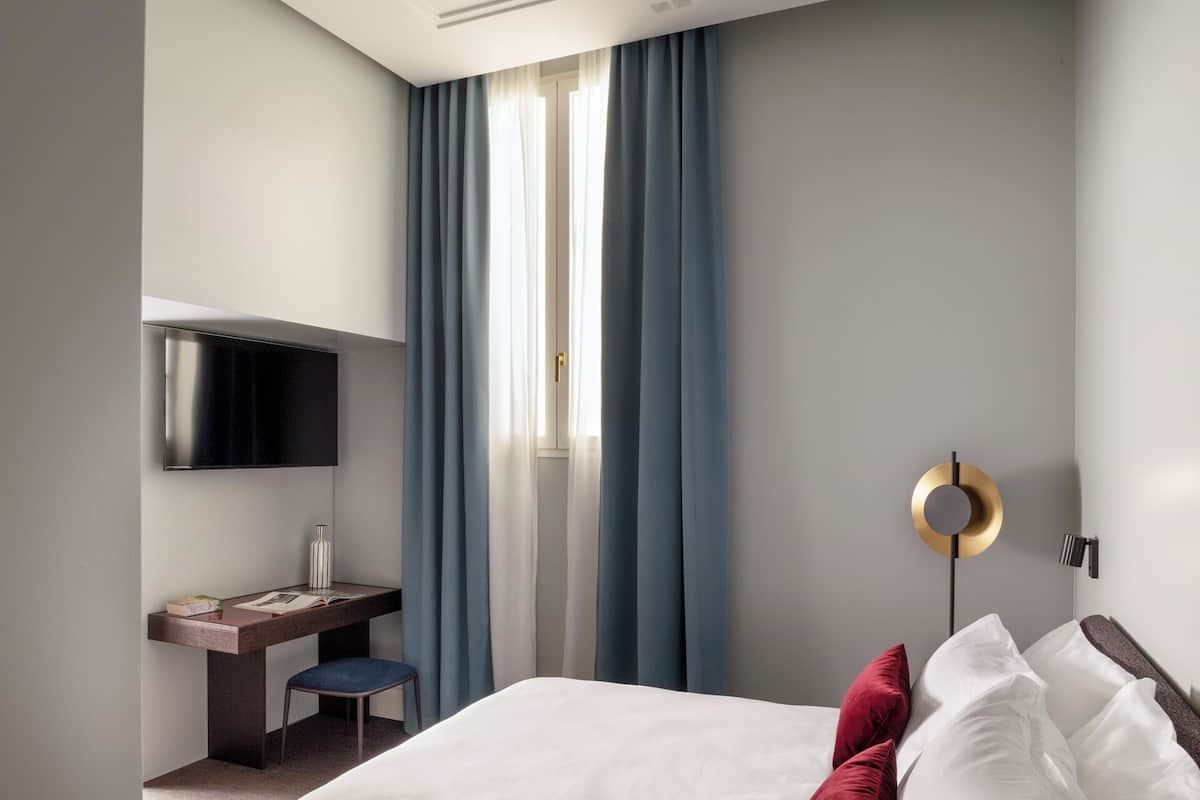 A modern hotel room with a bed, a desk with a chair, a television, and a wall-mounted light fixture.