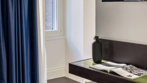 A modern hotel room with a large window, a flat-screen TV mounted on the wall, a bedside table with a book and a vase, and a green cushioned bench. - Golden House Vatican - Regolo (Rome)