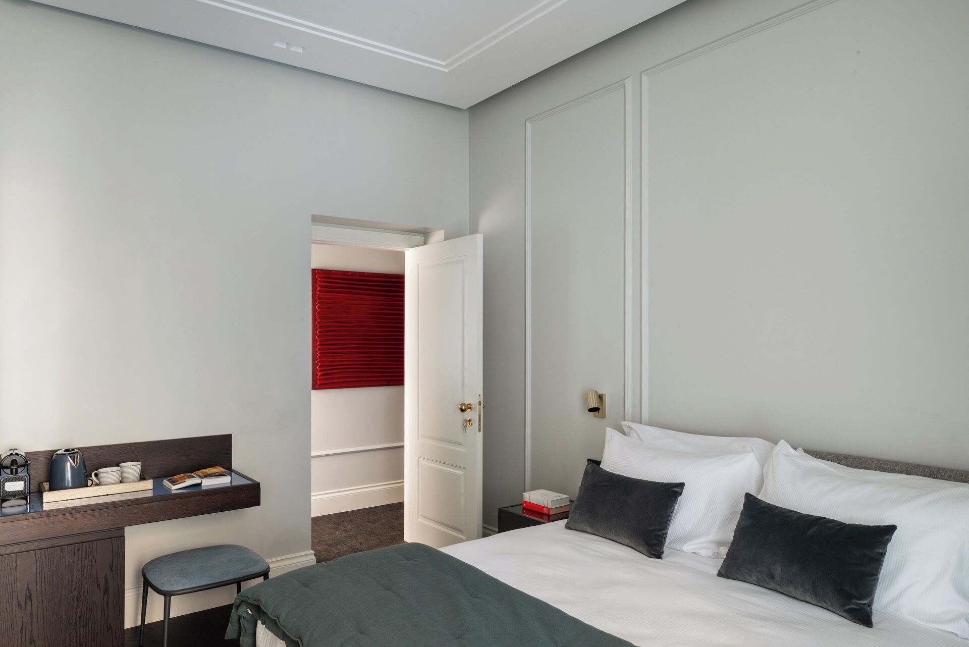 A hotel room with a bed, a desk with a chair, a small table with a kettle and cups, and a red curtain.
