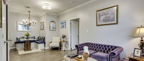 Una sala de estar con un sofá morado, una mesa de centro de vidrio y una mesita auxiliar de madera. Se observa una puerta blanca que da a otra habitación.