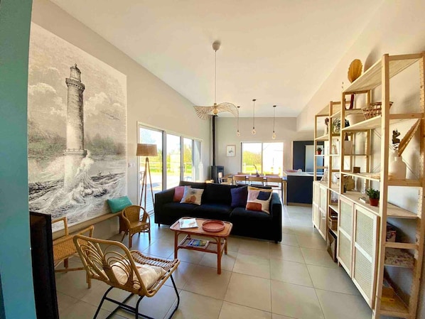Un salon spacieux, orné d’un grand tableau représentant un phare, avec un canapé aux couleurs sombres, des fauteuils en osier, une petite table et une bibliothèque en bois remplie d’objets.