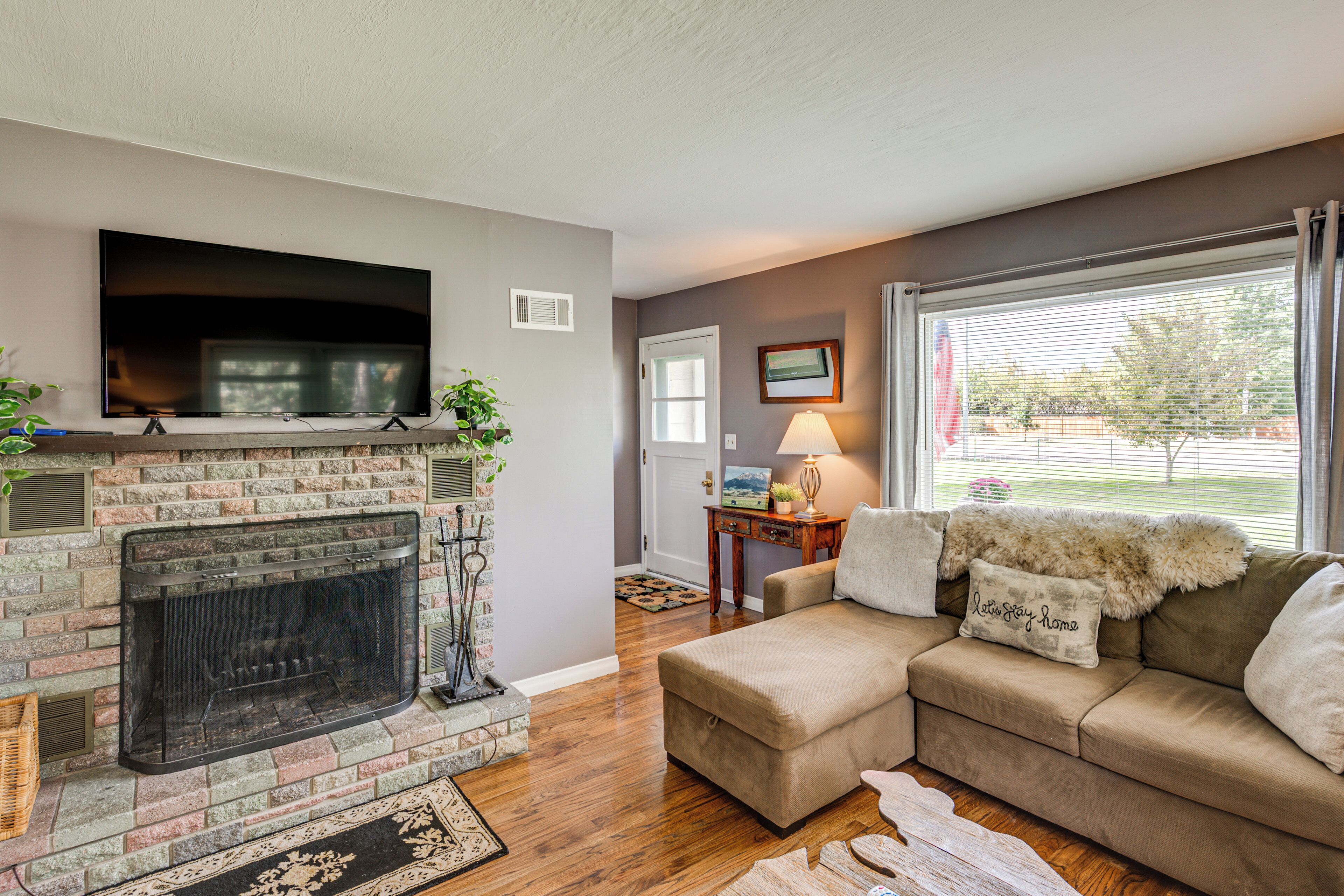 A living room with a fireplace, a television mounted above it, a sofa with decorative pillows, and a window with blinds.
