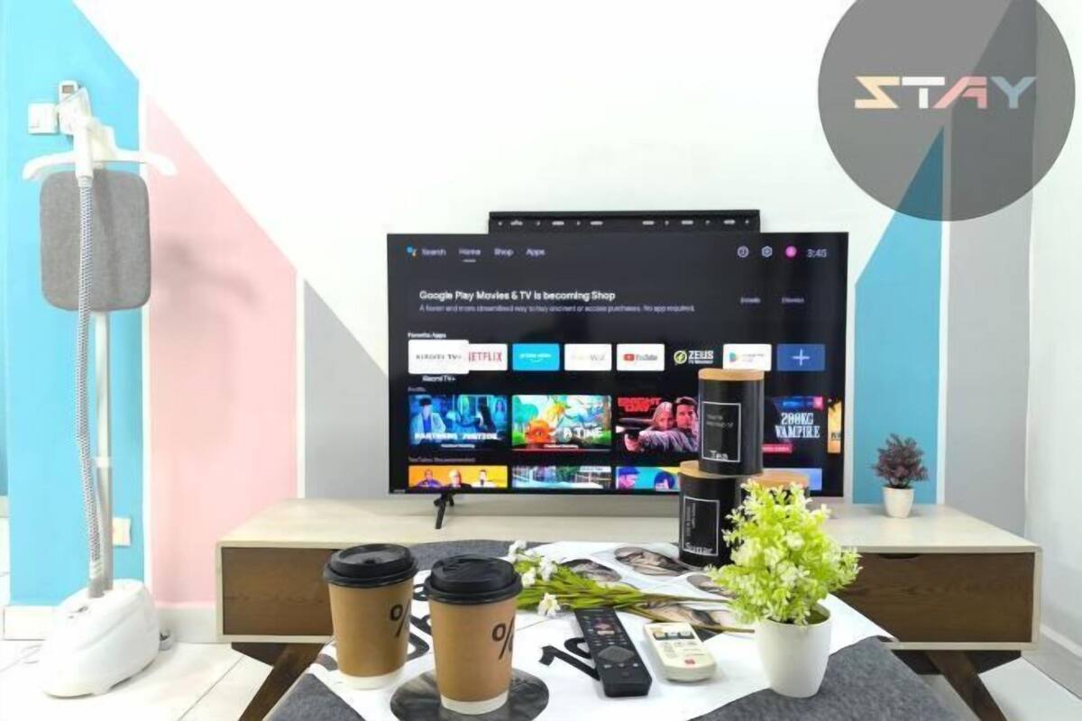 A modern living room with a flat-screen TV displaying a streaming service interface, a coffee table with drinks and remote controls, and a small plant.