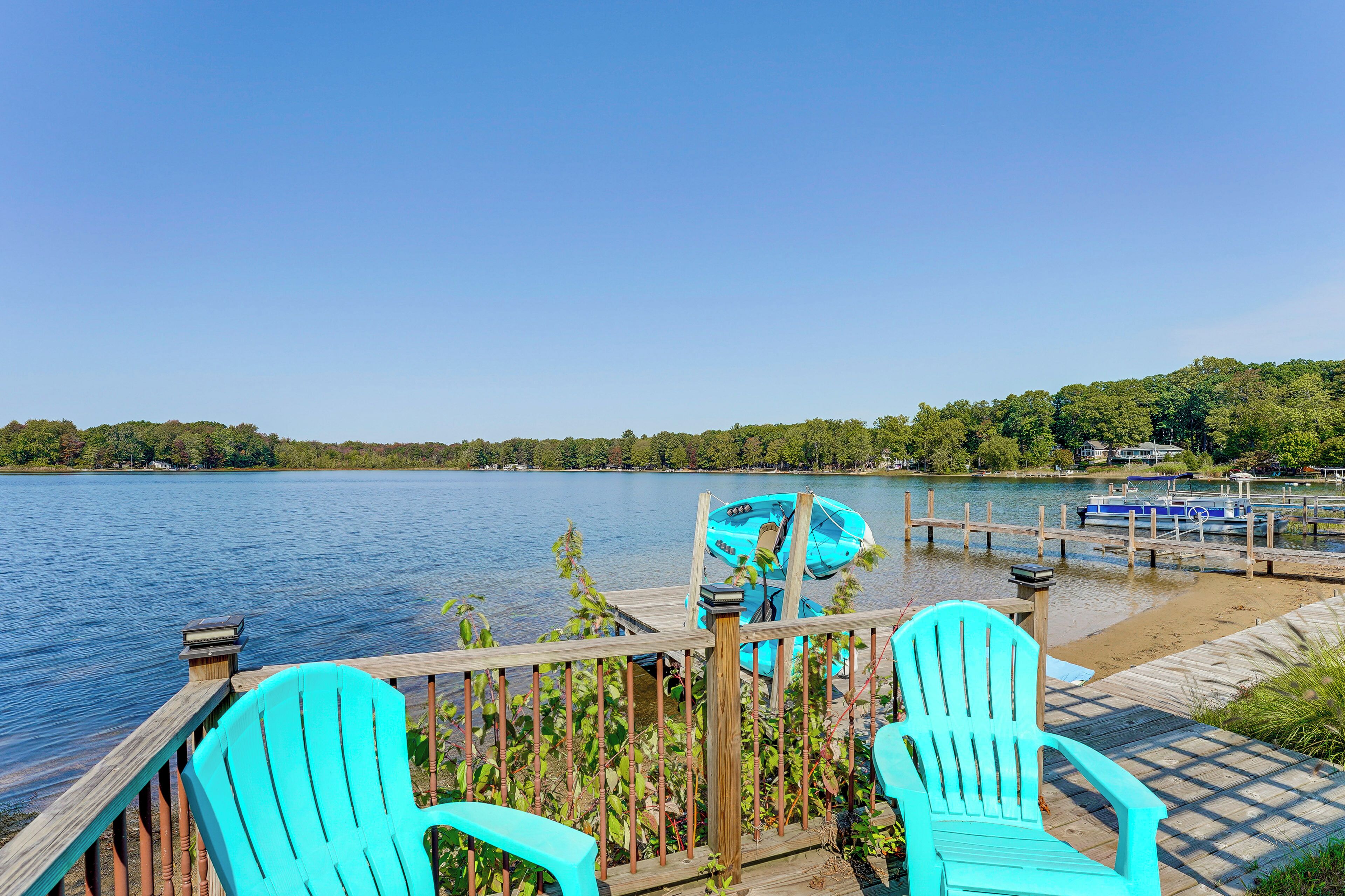 Une terrasse en bois avec des chaises turquoise donnant sur un lac paisible, un quai avec des bateaux et une rive bordée d’une forêt.