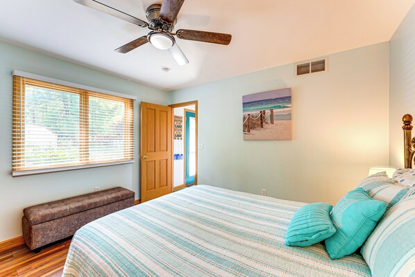 Un dormitorio con cama, un banco, un ventilador de techo y un cuadro con motivos playeros.