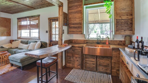 A rustic kitchen with wooden cabinets, a wooden countertop, and a built-in sink. There is a wooden dining table with benches and a green sofa in the living area.