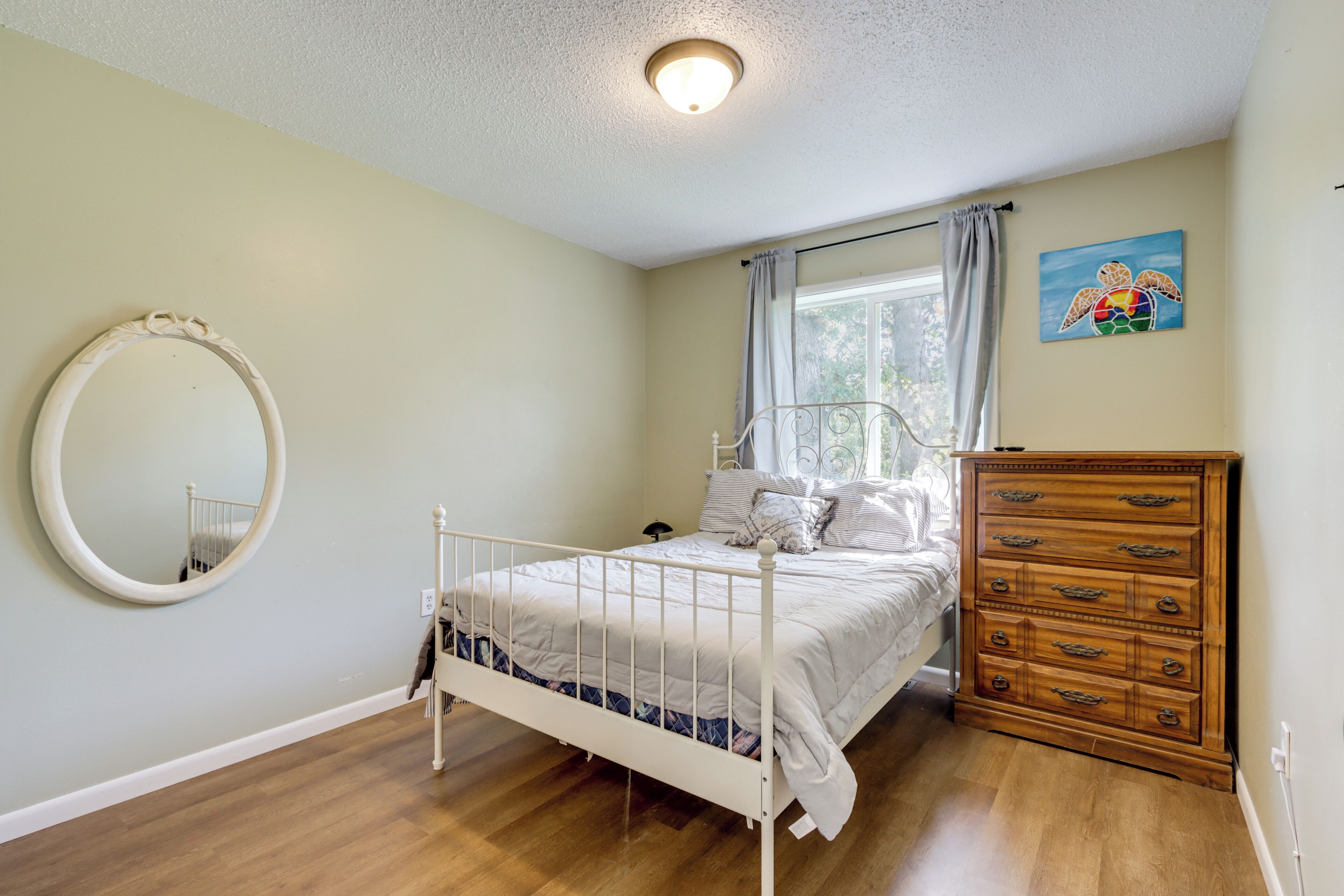 A bedroom with a white metal bed frame, a wooden dresser, a round mirror, and a window with curtains.