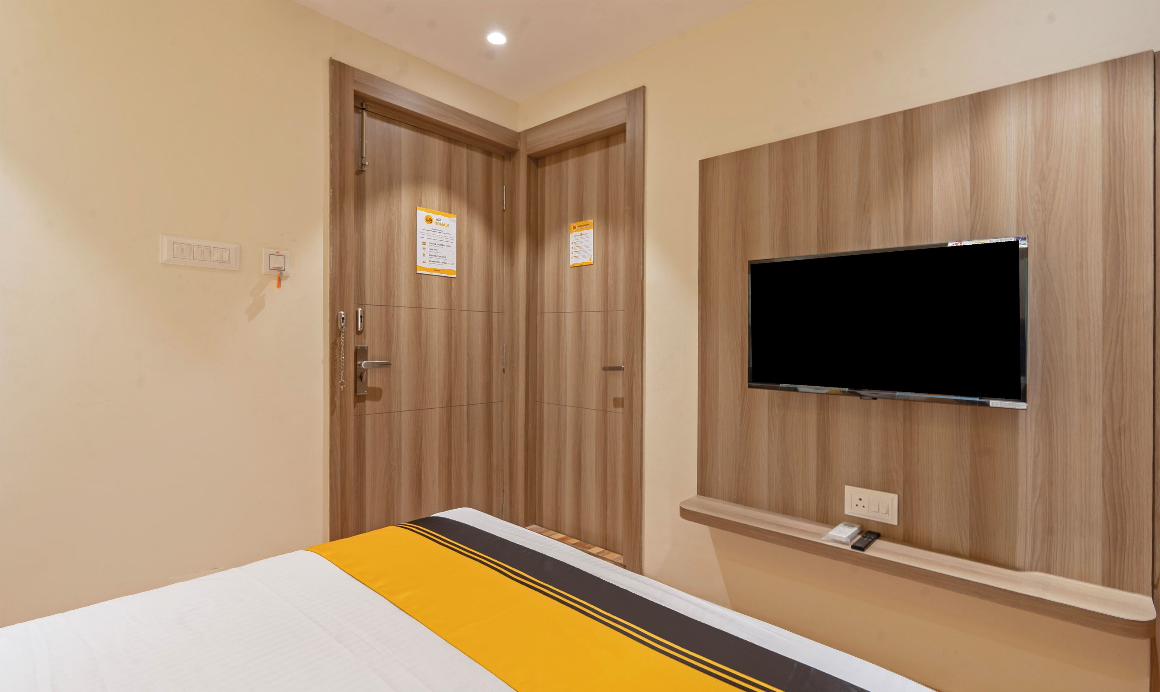 A hotel room with a bed, a TV mounted on the wall, and two doors with signs.