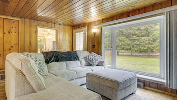 A wooden-paneled room with a sectional sofa, a window with a view of greenery, and a ceiling fan.