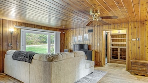 Ein gemütliches Wohnzimmer mit einem beigefarbenen Sofa, Holzwänden, einem Deckenventilator und einem großen Fenster mit Blick ins Freie.