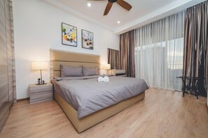 Une chambre moderne avec un grand lit, deux tables de chevet avec des lampes, un ventilateur de plafond et une vue sur l’extérieur à travers des rideaux légers.