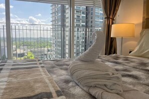 A hotel room with a large window, a bed with a striped blanket, a white pillow, and a lamp on a bedside table. - Encorp High Floor 1BR 3 pax Balcony Puteri Habour (Iskandar Puteri)