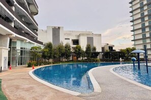 Une piscine à bord incurvé, entourée d’une allée pavée et jouxtant un immeuble à plusieurs étages doté de balcons.