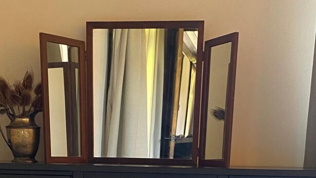 Una stanza con una scrivania in legno scuro, uno specchio, un vaso con fiori secchi e un letto bianco.
