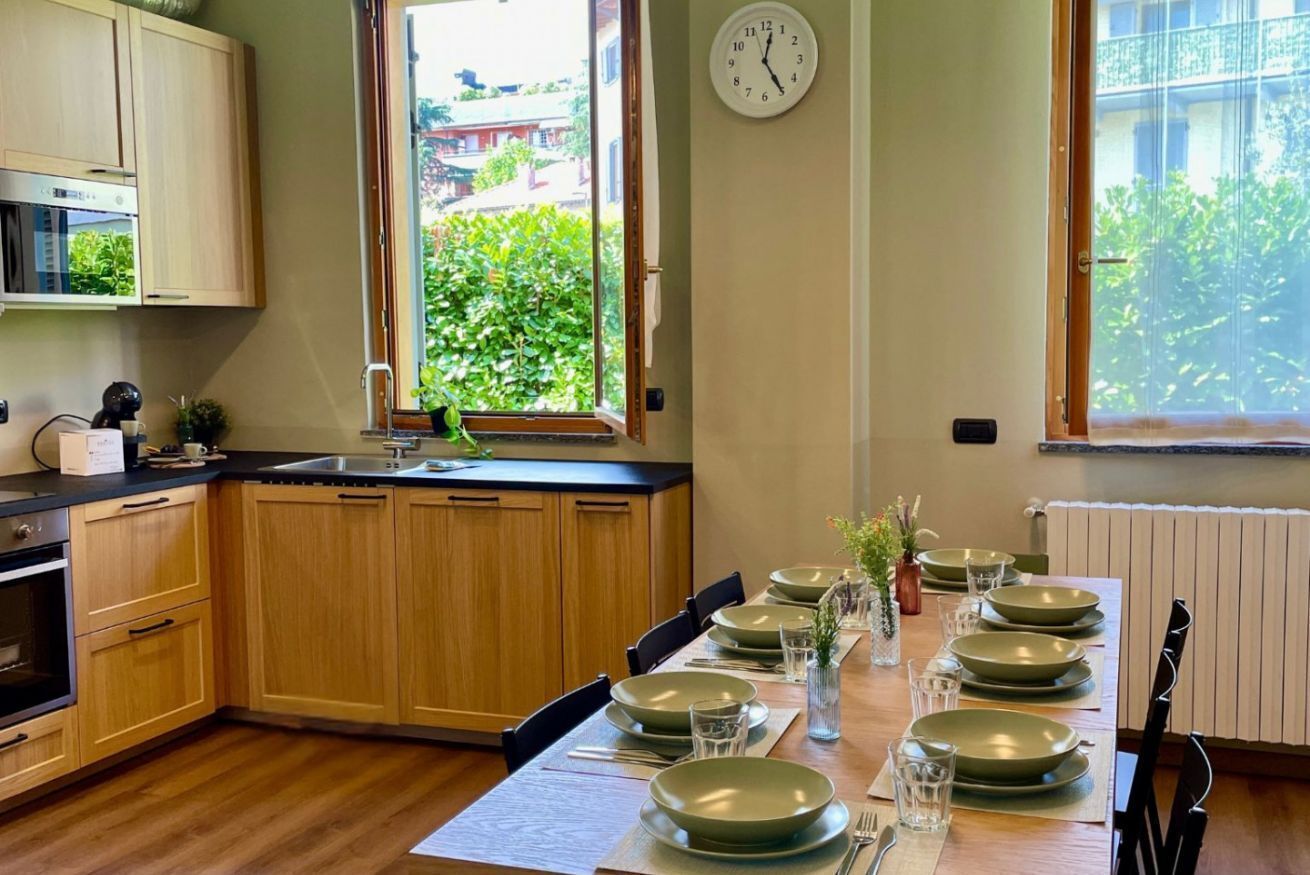 A kitchen with wooden cabinets, a window with a view of greenery, a clock on the wall, and a dining table set with plates and glasses.