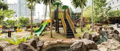 An outdoor playground with slides and stairs, surrounded by rocks and plants.