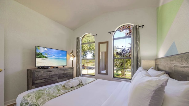Une chambre avec un grand lit, un téléviseur posé sur une commode en bois et une vue sur la plage, visible à travers des fenêtres en arc de cercle.