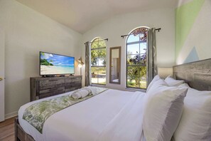 A bedroom with a large bed, a television on a wooden dresser, and a view of a beach through arched windows.